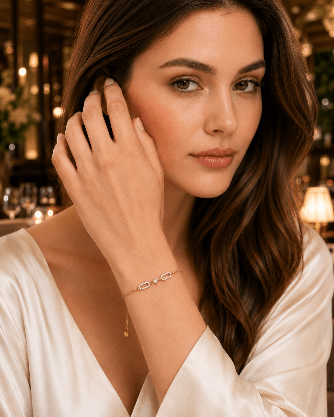 A woman wearing a delicate gold chain bracelet with a small oval link and a dangling charm, posed in a warm, softly lit indoor setting.