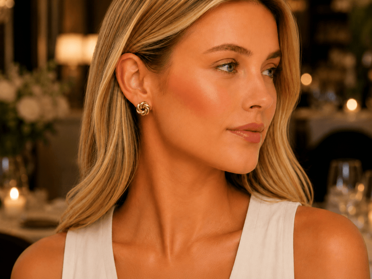 A close-up lifestyle shot of a woman with blonde hair wearing a small gold spiral stud earring in a warmly lit restaurant setting.