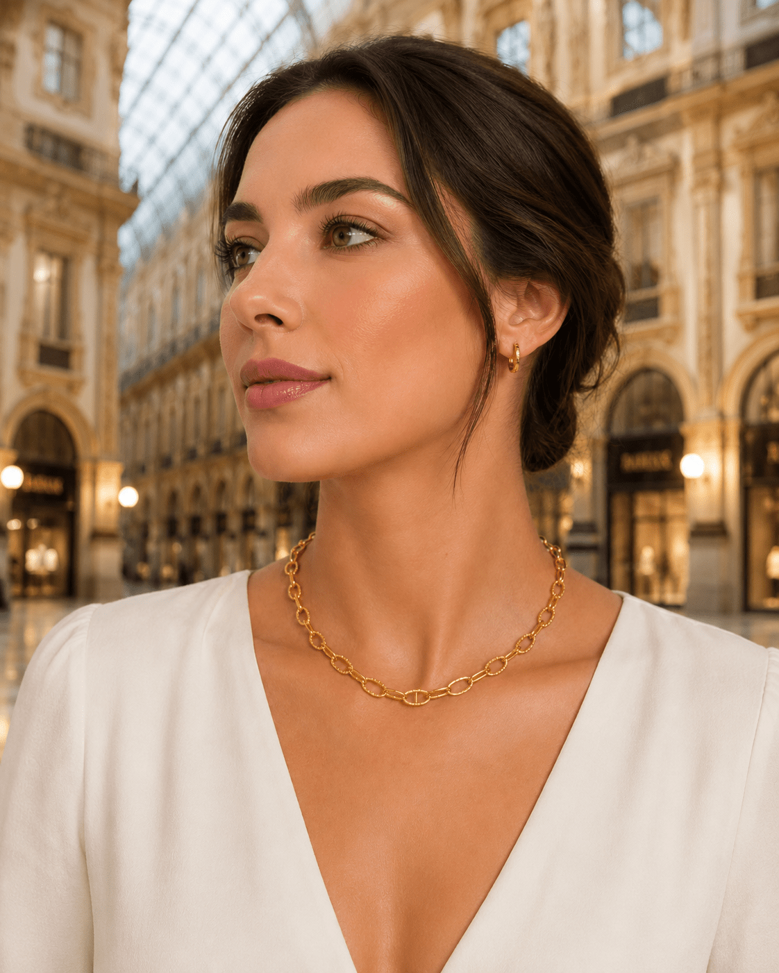 A woman wearing a delicate gold chain necklace and a small gold hoop earring, photographed in a grand indoor shopping arcade with warm lighting and architectural detail in the background.