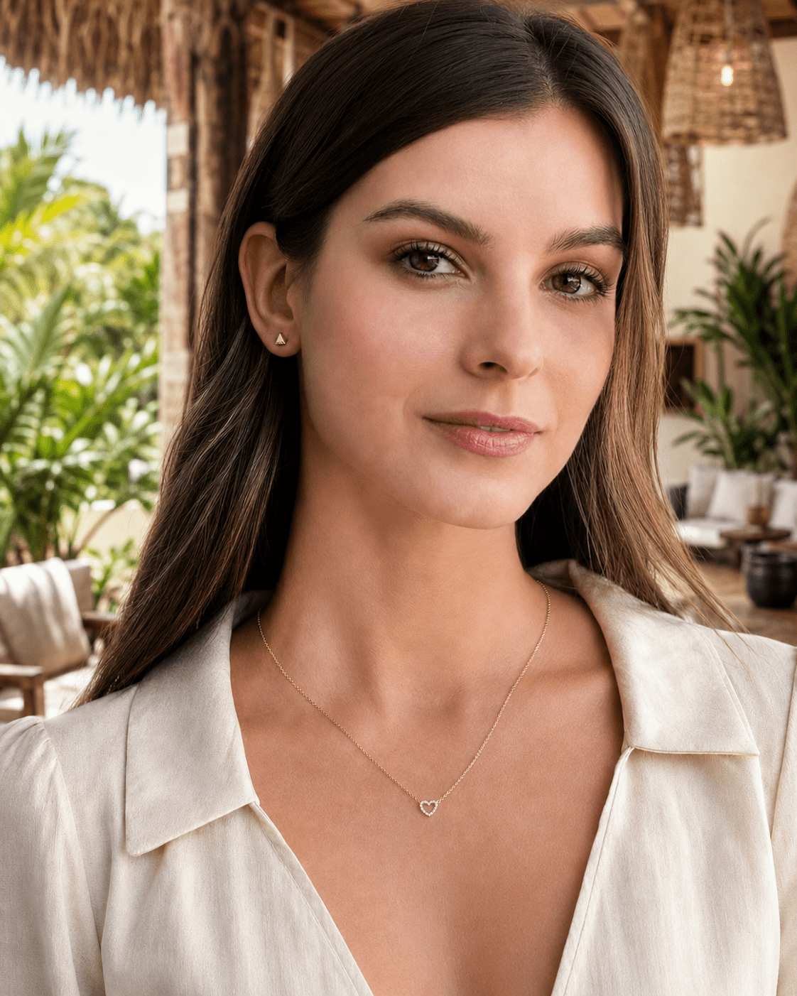 A woman wearing a delicate gold heart pendant necklace and a small stud earring, photographed in a sunny outdoor cabana setting.