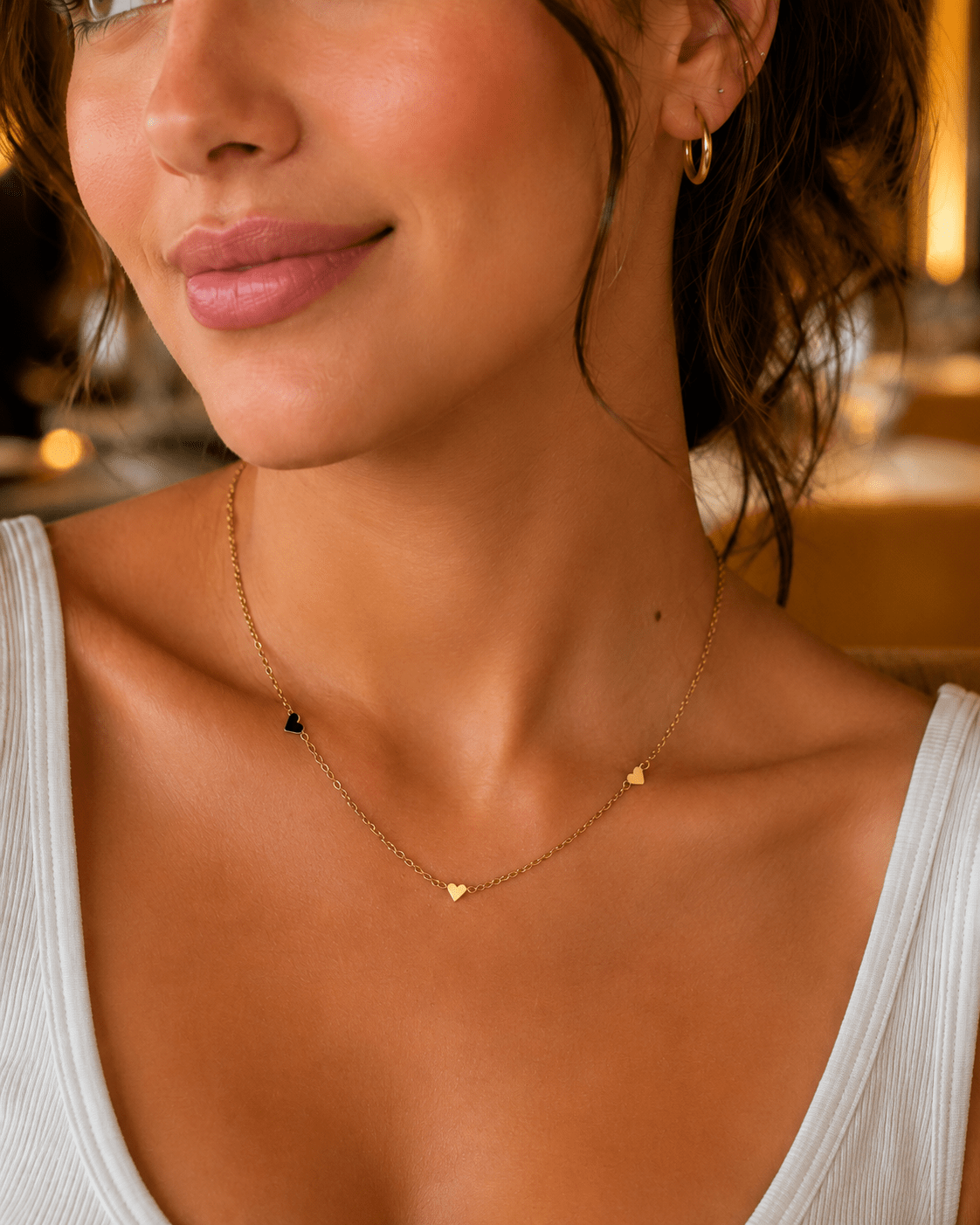 A close-up lifestyle shot of a woman wearing a delicate gold chain necklace with three small gold heart charms, styled with a white ribbed tank top.