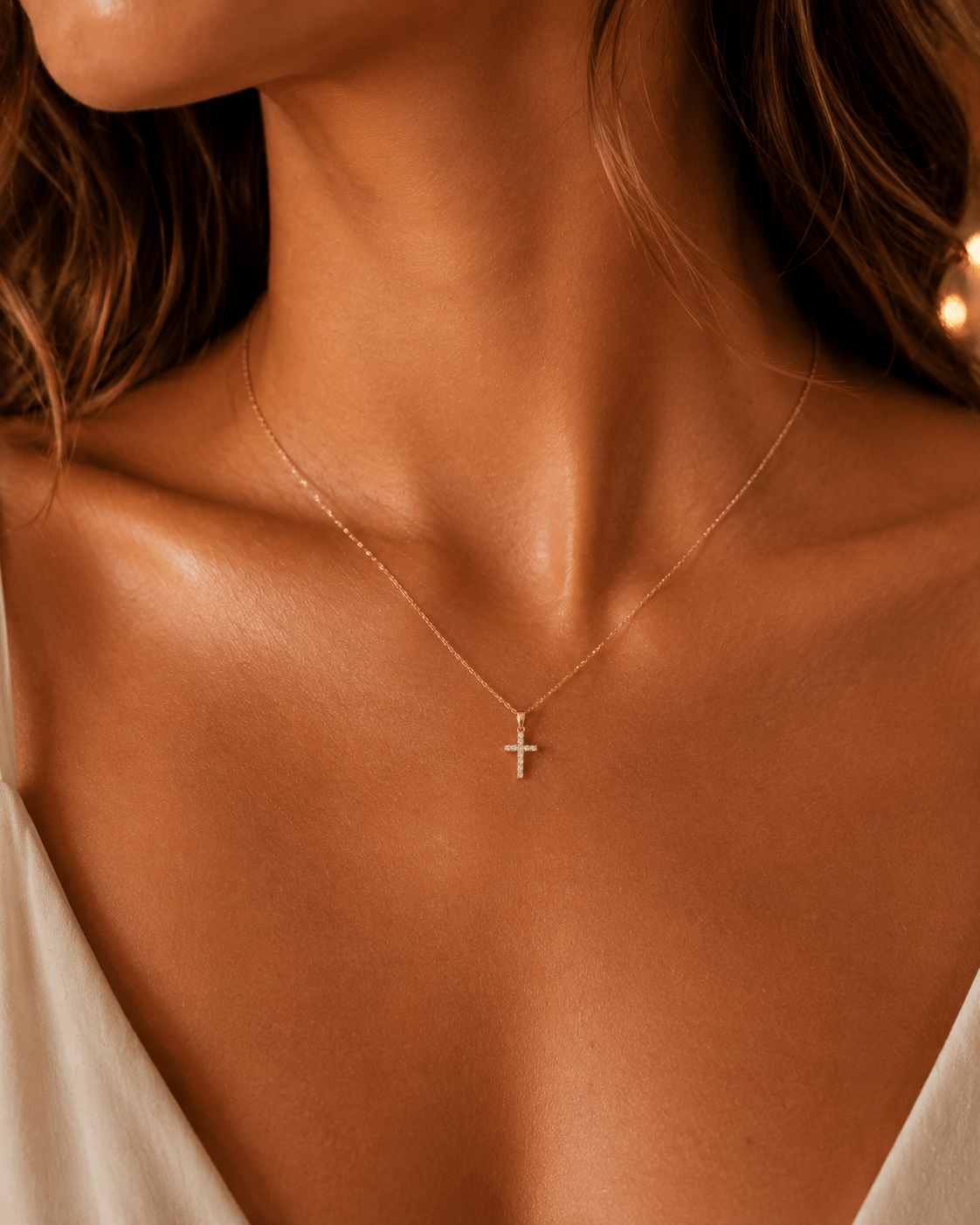 A delicate gold cross pendant necklace worn by a model, shown in a close-up shot highlighting the jewelry against warm skin tones.