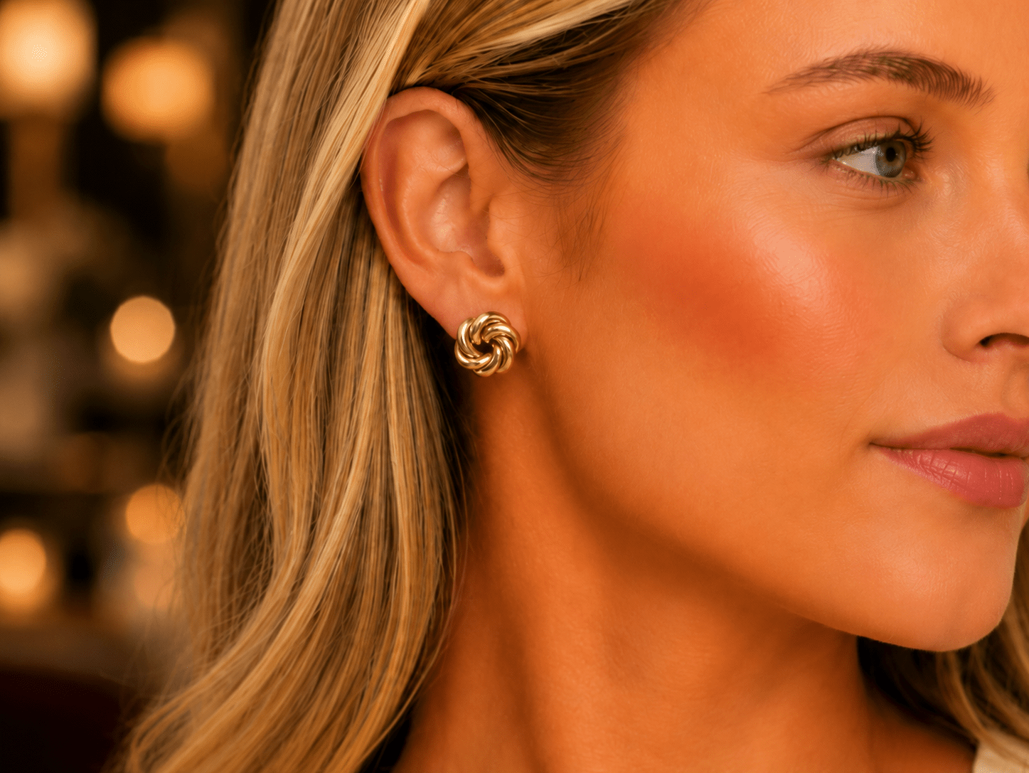 A close-up of a woman's ear wearing a small gold spiral stud earring, with warm blurred lights in the background.