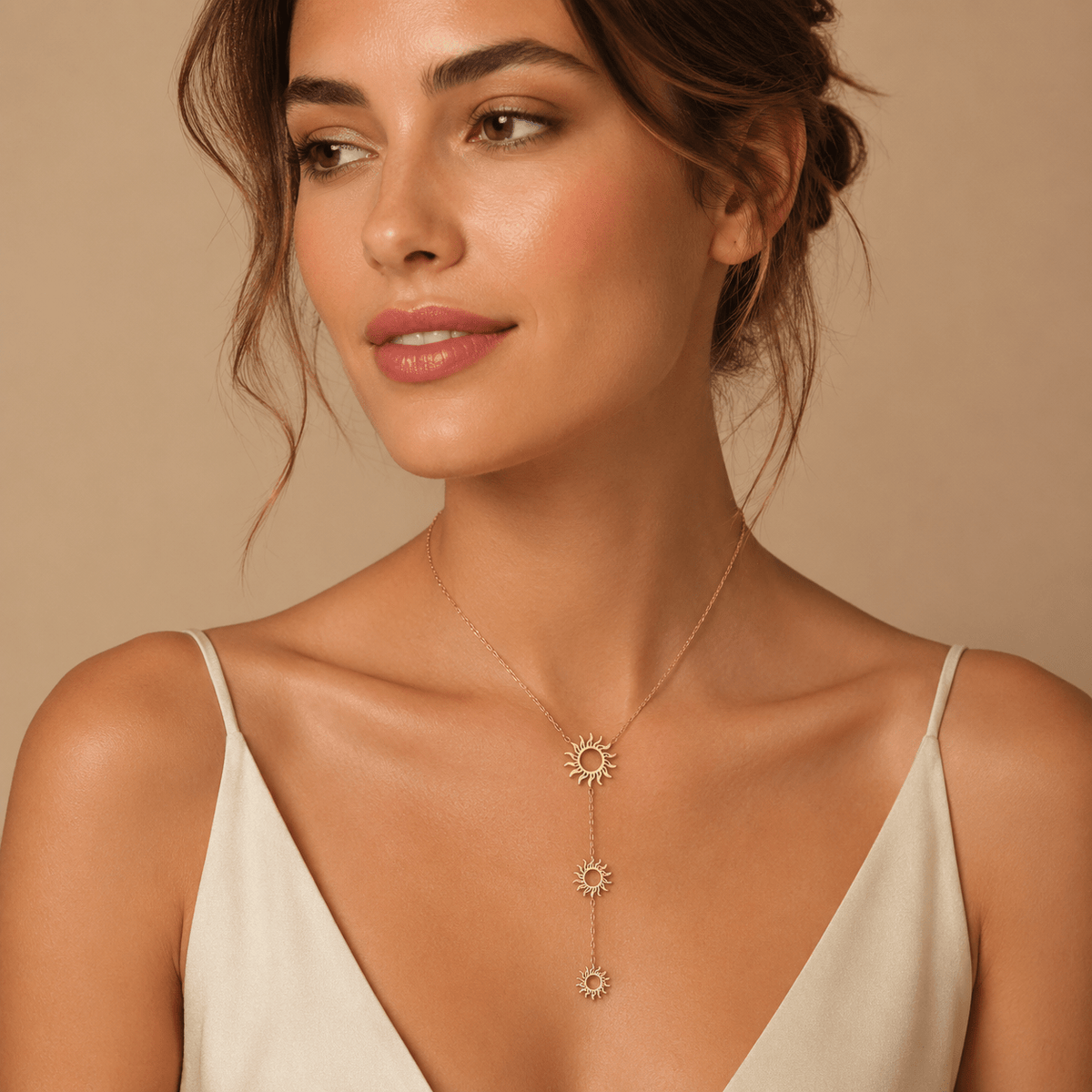 A woman wearing a delicate gold lariat-style necklace with three sunburst charms, styled with a cream-colored spaghetti strap top against a warm beige background.