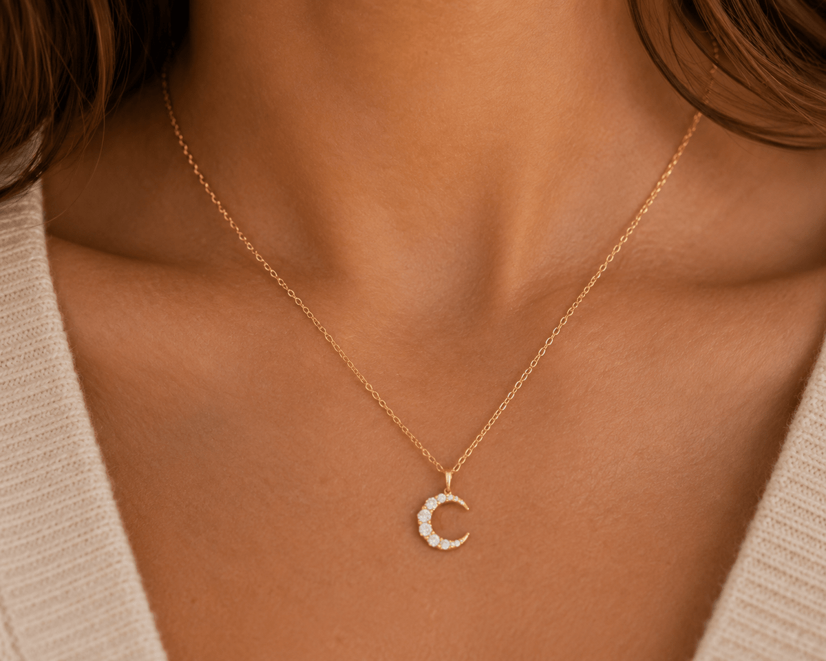 A close-up of a model wearing a delicate gold chain necklace with a crescent moon pendant studded with small clear crystals, styled with a beige knit top.