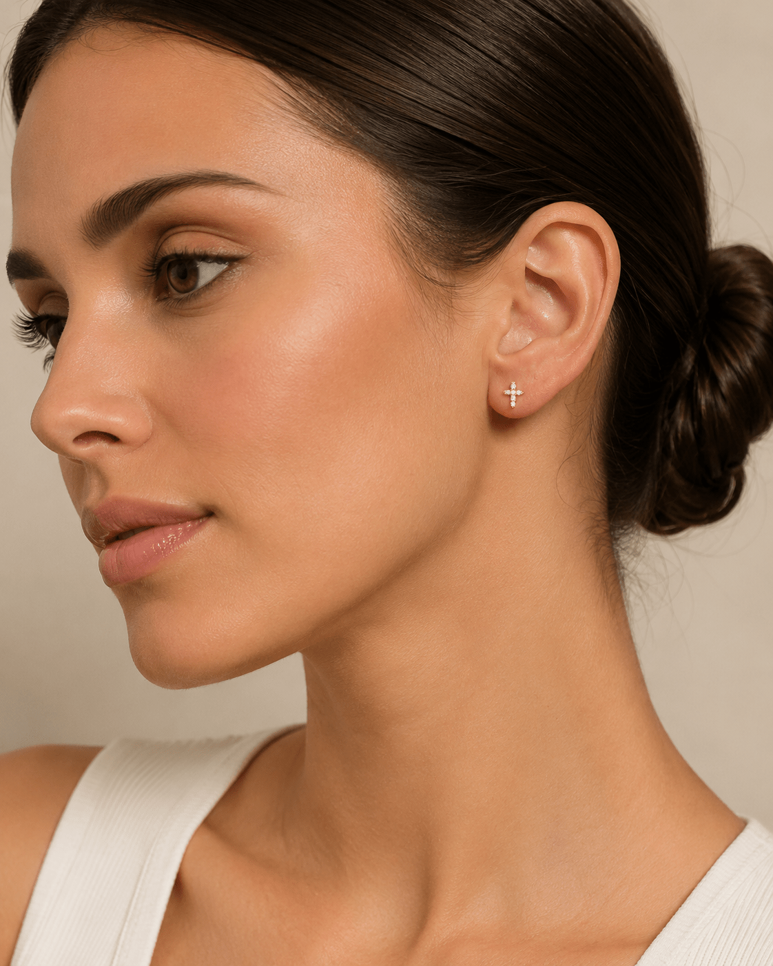 A close-up of a woman wearing a delicate gold cross stud earring, shown in profile against a soft beige background.