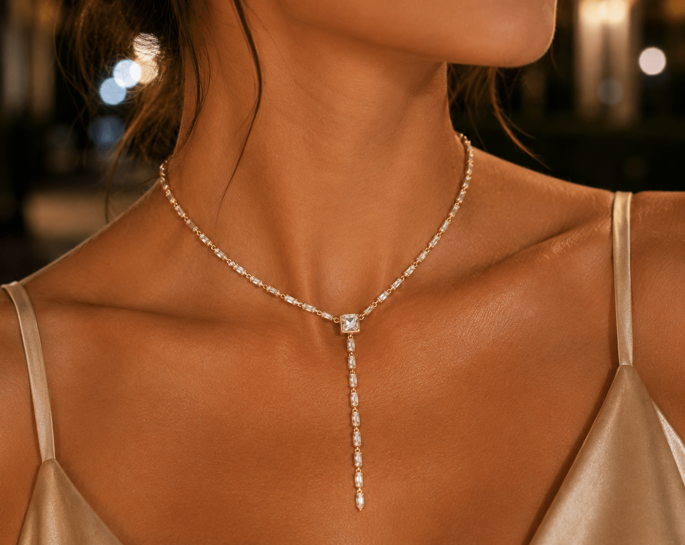 A close-up of a woman wearing a delicate CZ simulated diamond Y-necklace with a central square-cut CZ pendant and a long drop chain, styled with a satin slip dress against a softly blurred night background.