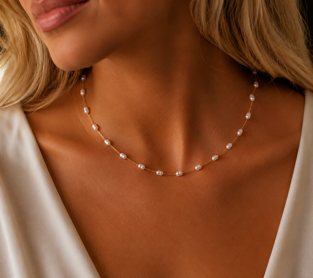 A close-up of a woman wearing a delicate gold necklace with small white pearls, styled with a white satin top.