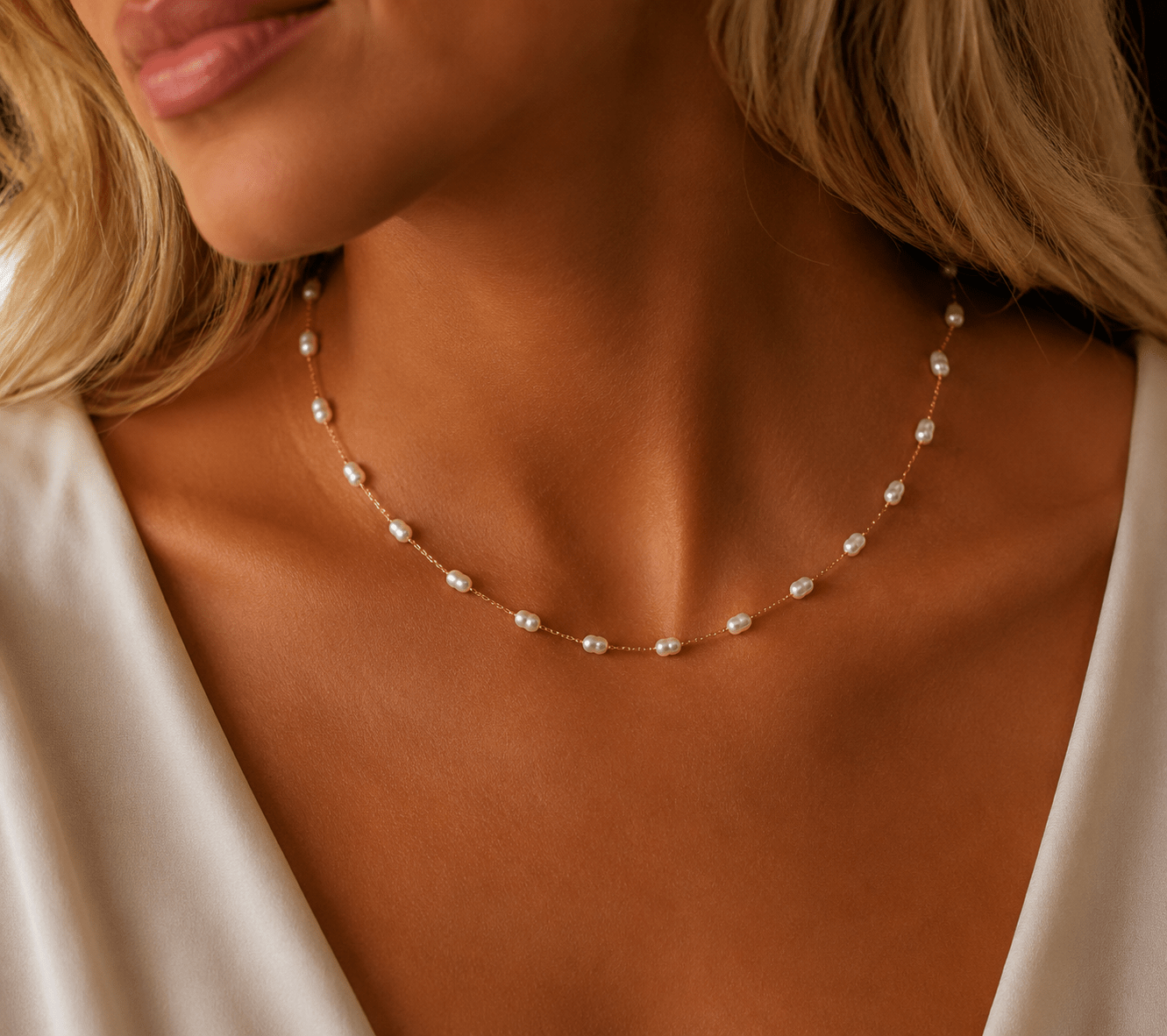 A close-up of a woman wearing a delicate gold necklace with small white pearls, styled with a white satin top.