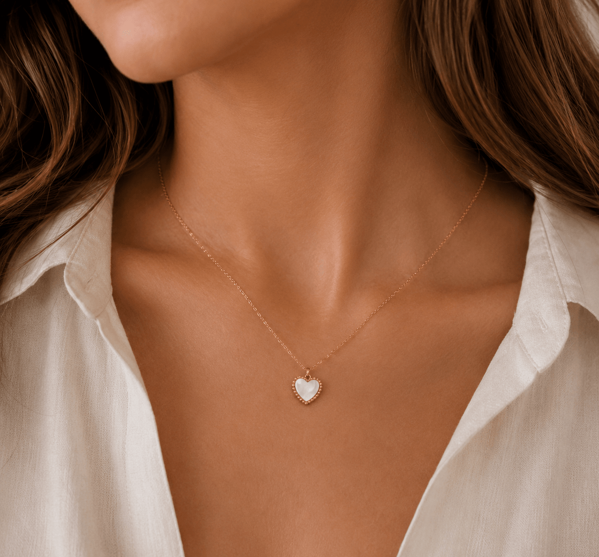 A close-up of a woman wearing a delicate gold chain necklace with a small white heart-shaped pendant, styled with a white collared shirt.
