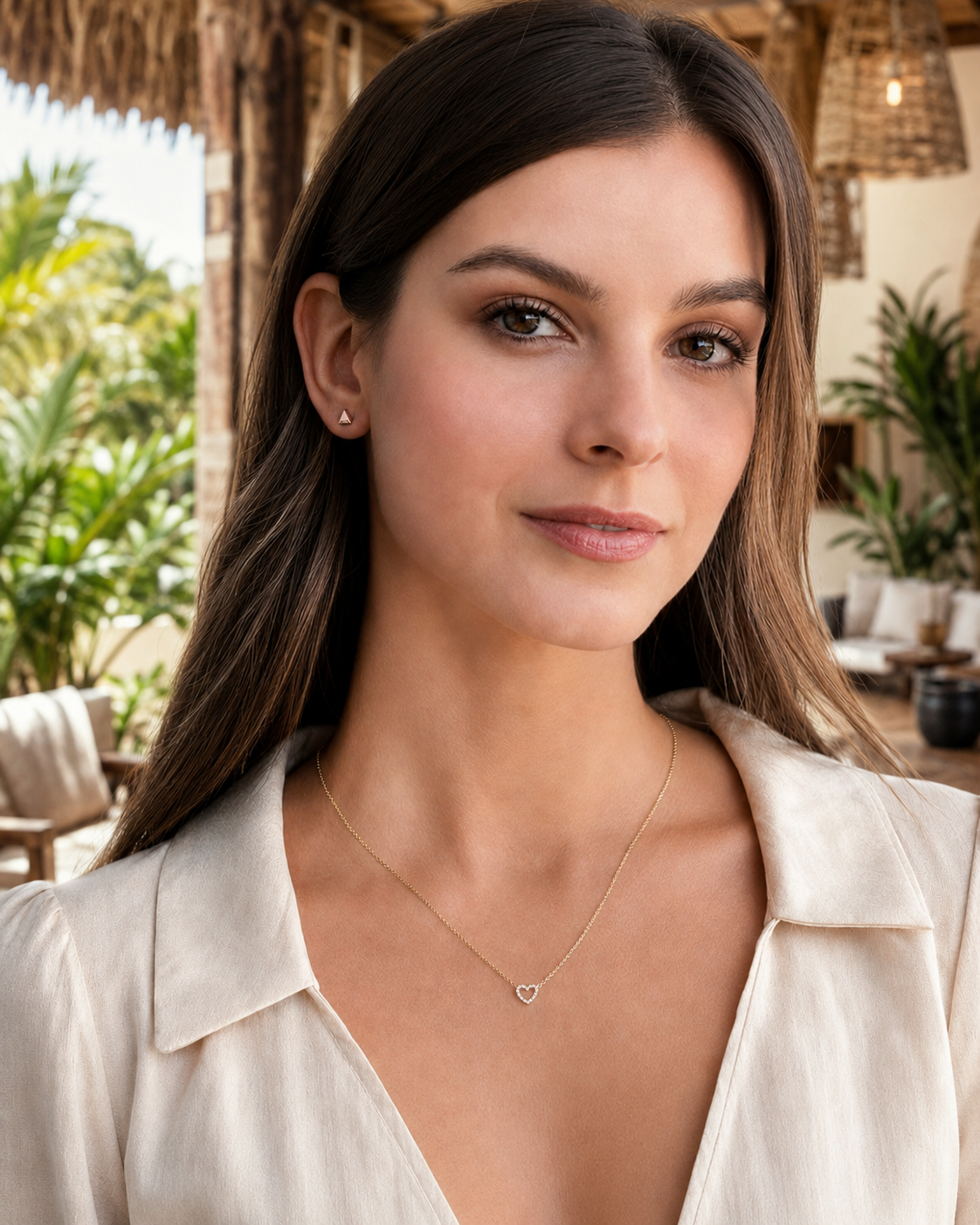 A woman wearing a delicate gold heart pendant necklace and a small stud earring, photographed in a sunny outdoor cabana setting.