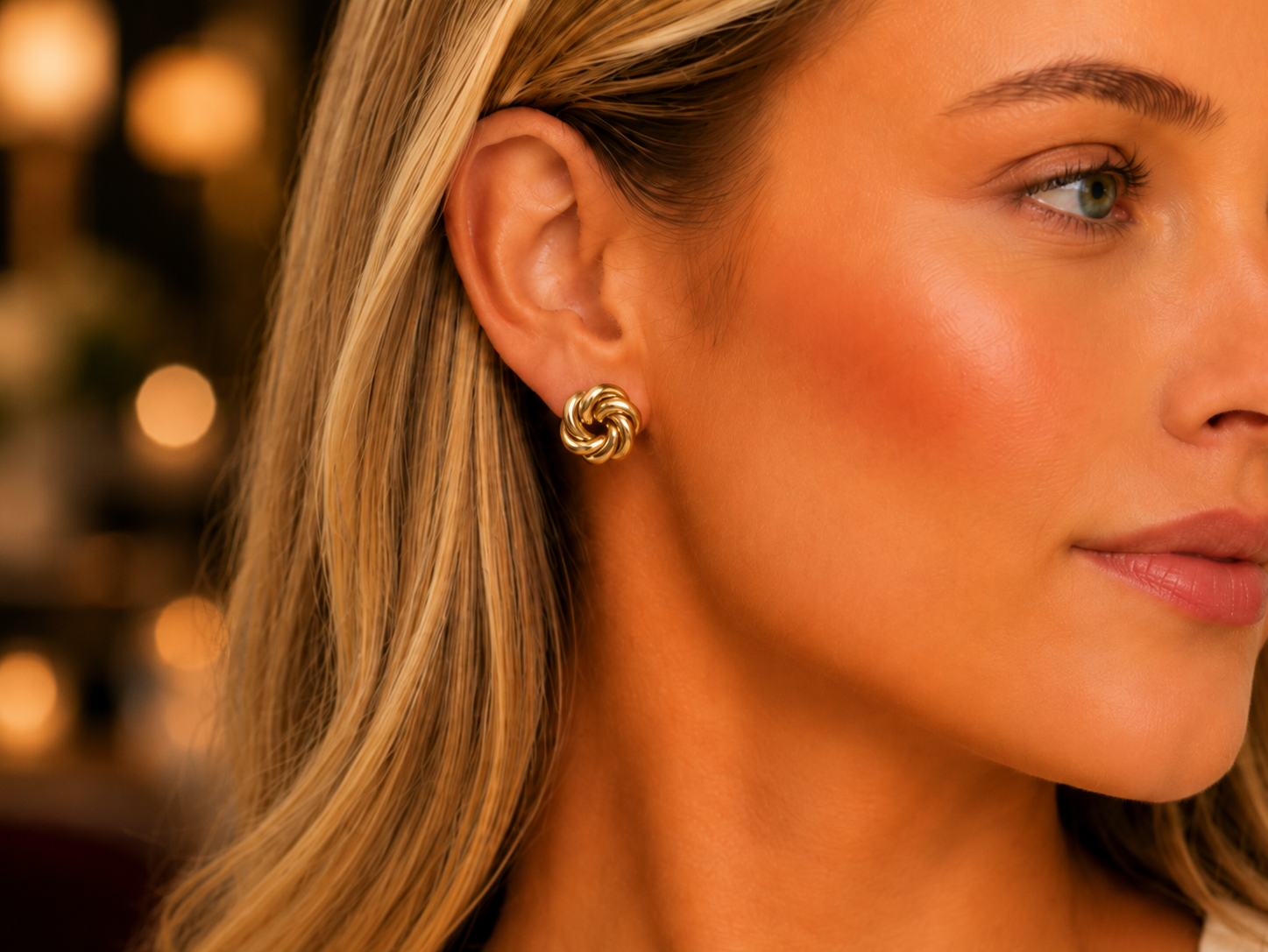 A close-up of a woman's ear wearing a small gold spiral stud earring, with warm blurred lights in the background.