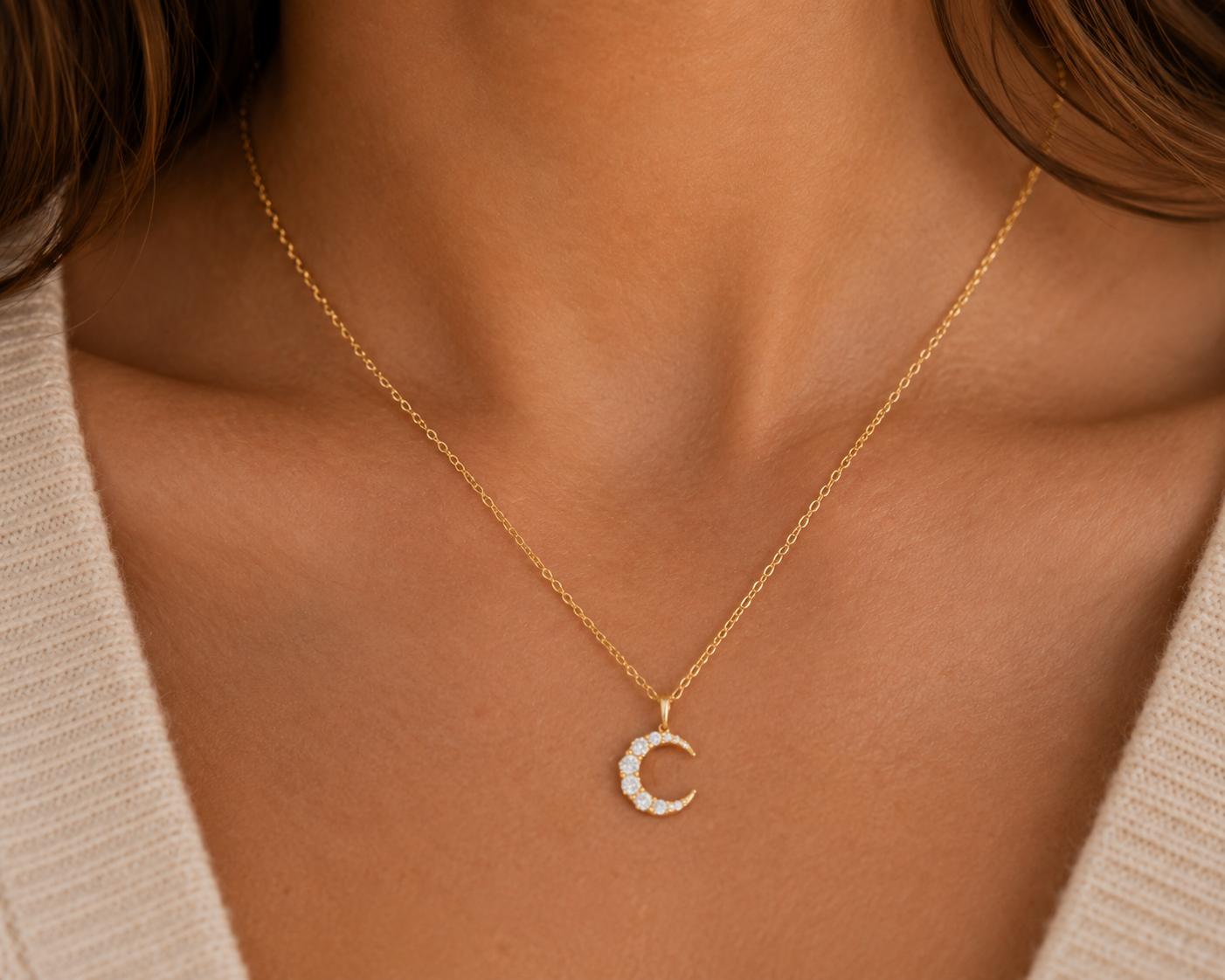 A close-up of a model wearing a delicate gold chain necklace with a crescent moon pendant studded with small clear crystals, styled with a beige knit top.