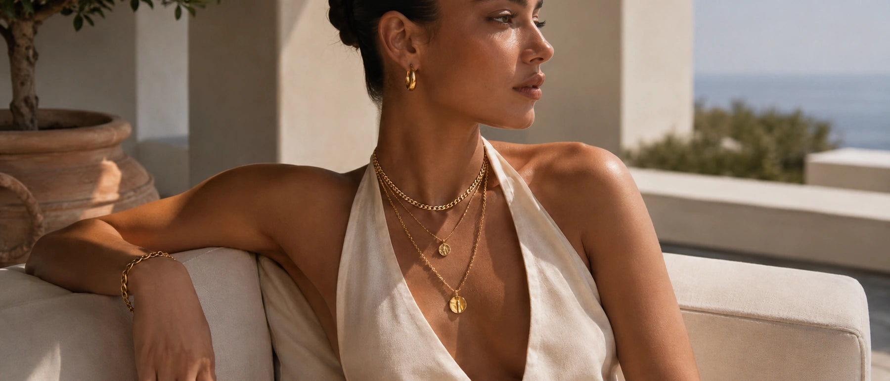 Woman wearing layered gold pendant necklaces, hoop earrings and a chain bracelet on a seaside terrace