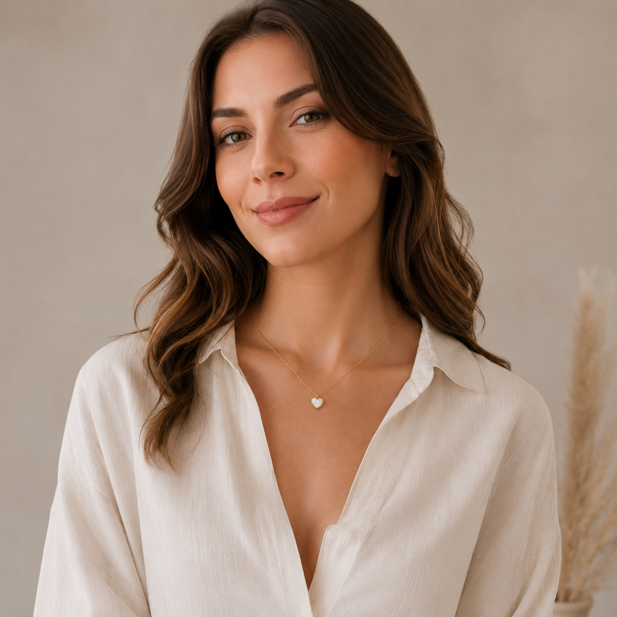 A woman with wavy brown hair wearing a cream-colored linen button-down shirt and a delicate gold heart pendant necklace, posing against a soft beige background.